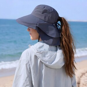 May include: A dark grey sun hat with a wide brim and a neck flap, designed for sun protection. The hat has a ponytail opening at the back. The person is wearing a light grey jacket, with the sea and beach in the background.