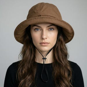 May include: A solid brown bucket hat with a wide, downward-sloping brim and a structured crown. The hat features a thin black adjustable chin strap with a plastic toggle cord lock, providing a secure fit around the head and neck area.
