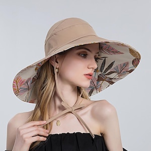 May include: A beige sun hat with a wide brim, featuring a floral pattern on the underside. The hat has a matching chin strap and is designed for sun protection. The brim's interior displays a colorful botanical print.