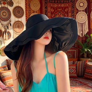 Puede incluir: Un sombrero negro para el sol con un ala ancha y ondulada. El sombrero lo lleva una persona que lleva un top halter turquesa. El fondo presenta tapices y cojines decorativos.