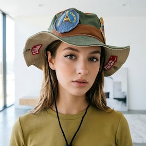 Puede incluir: Un sombrero de pescador verde, marrón y beige con varios parches. El sombrero tiene un ala ancha y una correa de barbilla negra. Los parches incluyen un diseño azul y amarillo y un parche rojo con texto blanco. El sombrero lo lleva una persona.