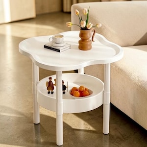 Op de afbeelding: Een witte, tweelaagse bijzettafel met een uniek, wolkenachtig tafelblad. De tafel heeft een onderste plank en wordt ondersteund door vier witte poten. Een kleine bruine vaas met bloemen, een kopje en een boek staan ​​bovenop. De onderste plank bevat een fruitschaal en decoratieve beeldjes.