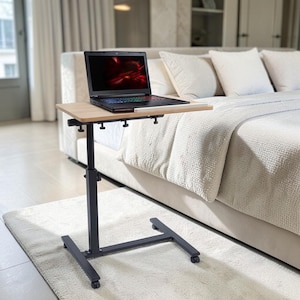 May include: A black, adjustable laptop table with a light wood-colored surface. The table has a black base with wheels and is positioned over a white rug. A laptop with a lit screen sits on the table.