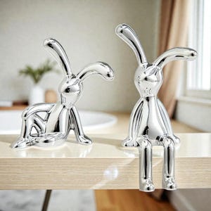 May include: Two silver bunny figurines. One is posed sitting, the other is sitting with its legs dangling. The bunnies have long ears and are made of a shiny, reflective material. They are placed on a light-colored wooden surface.