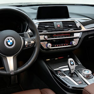 May include: Interior of a BMW car, showing a black leather steering wheel with a crystal-encrusted emblem. The dashboard has a central display and crystal-decorated control knobs. The gear shift and other interior elements are also embellished with crystals.