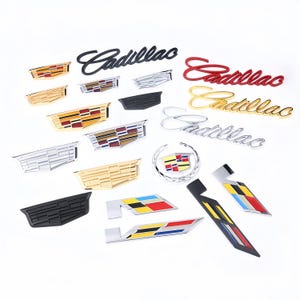 May include: A selection of Cadillac emblems and logos in diverse colours and designs. The image shows the word "Cadillac" in black, red, and gold, alongside wing-shaped emblems in silver, gold, and black, with colourful geometric patterns.