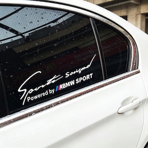 以下が含まれることがあります： 白い車の窓用ステッカー。スタイリッシュなフォントで「Sports Souizmd」と書かれています。その下に「Powered by」と「ABMW SPORT」の文字が表示されています。ステッカーは、シルバーのトリムが付いた白い車に貼られています。