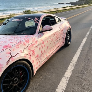 May include: A white sports car with a cherry blossom design parked on a coastal road. The car has black wheels with blue accents and a black roof. The car's body is covered in pink cherry blossom flowers and branches.