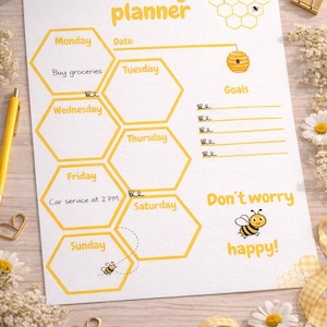 May include: A white weekly planner with yellow honeycomb-shaped sections for each day of the week. The planner includes the text "Weekly planner", "Don't worry, happy!", and a bee graphic. It also has space for goals and a date.