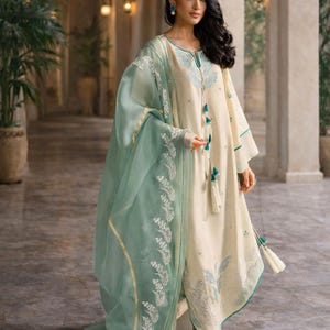 May include: Cream-colored embroidered tunic and matching pants with a sheer, mint green dupatta. The outfit features intricate teal and white floral embroidery and tassel details. The ensemble is a traditional South Asian style.