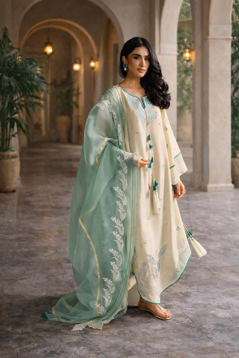 May include: Cream-colored embroidered tunic and matching pants with a sheer, mint green dupatta. The outfit features intricate teal and white floral embroidery and tassel details. The ensemble is a traditional South Asian style.