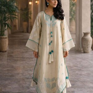 May include: Cream-colored long dress with turquoise embroidery and tassel details. The dress features wide sleeves and a matching trim. The outfit includes white pants and sandals. The style is elegant and suitable for special occasions.