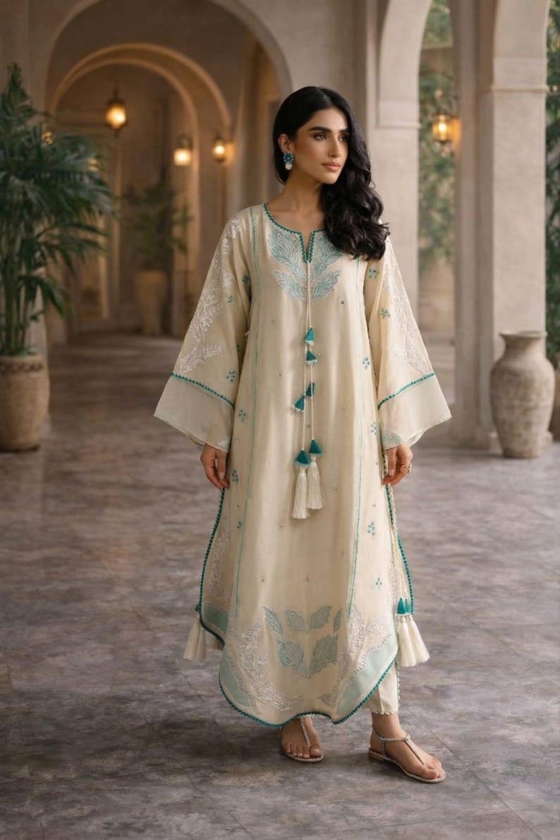May include: Cream-colored long dress with turquoise embroidery and tassel details. The dress features wide sleeves and a matching trim. The outfit includes white pants and sandals. The style is elegant and suitable for special occasions.