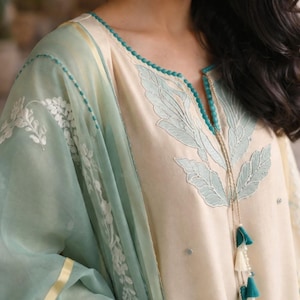May include: Cream-colored tunic with light blue leaf embroidery and a matching sheer scarf. The tunic features a beaded neckline and tassel details. The sleeves and scarf have white floral embroidery and a gold trim.