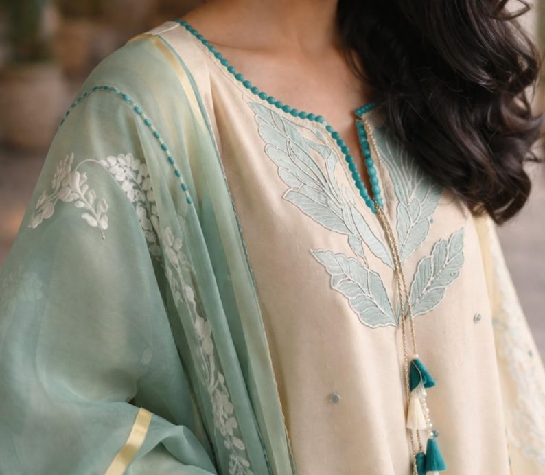 May include: Cream-colored tunic with light blue leaf embroidery and a matching sheer scarf. The tunic features a beaded neckline and tassel details. The sleeves and scarf have white floral embroidery and a gold trim.