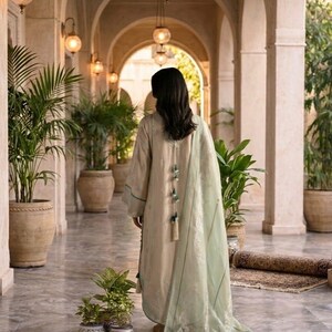 May include: A woman wearing a beige dress with green accents and a matching shawl, walking down a marble hallway. The dress features decorative tassels. The hallway is lined with potted plants and arched doorways, illuminated by hanging globe lights.