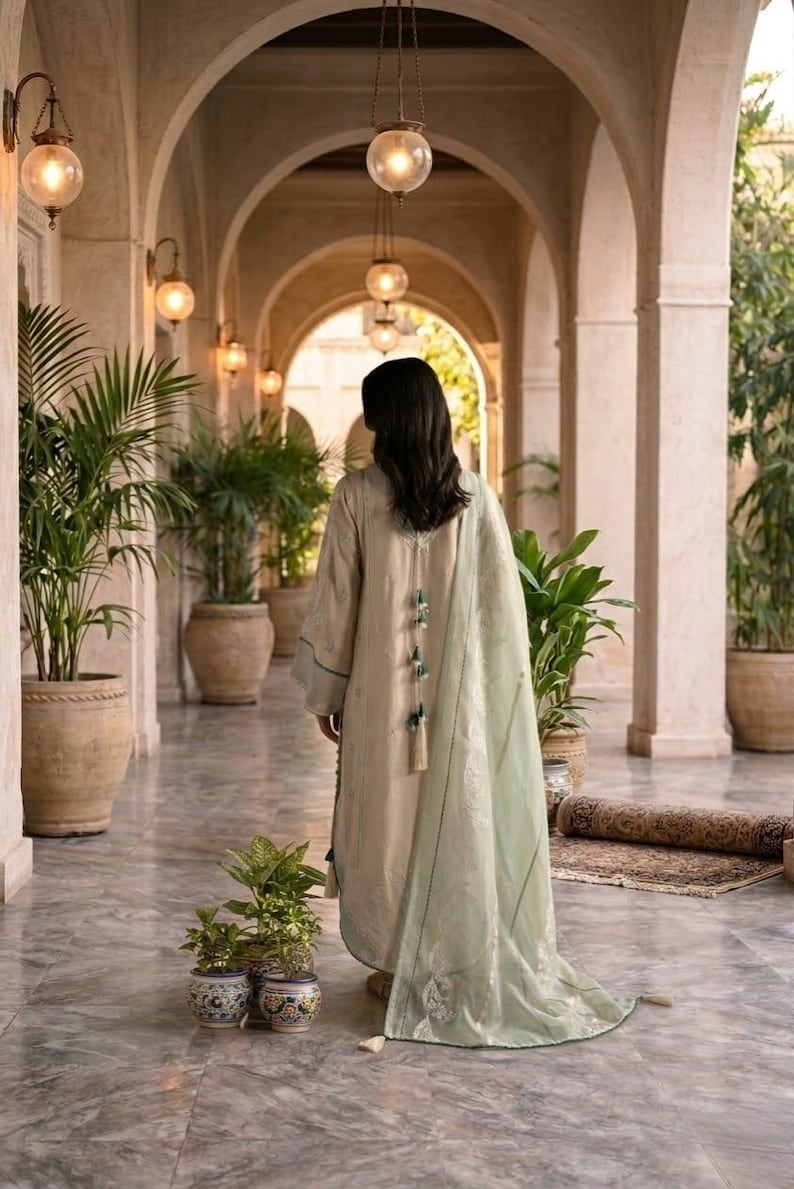 May include: A woman wearing a beige dress with green accents and a matching shawl, walking down a marble hallway. The dress features decorative tassels. The hallway is lined with potted plants and arched doorways, illuminated by hanging globe lights.