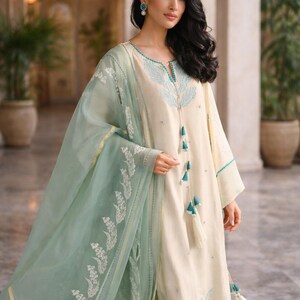 May include: Cream-colored tunic and matching pants with turquoise embroidery and tassels. A sheer, mint green scarf with white floral embroidery drapes over the outfit. The ensemble is elegant and suitable for special occasions.