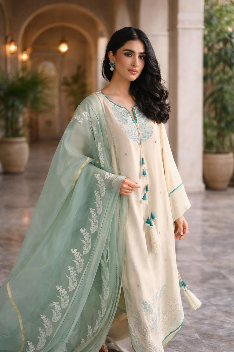 May include: Cream-colored tunic and matching pants with turquoise embroidery and tassels. A sheer, mint green scarf with white floral embroidery drapes over the outfit. The ensemble is elegant and suitable for special occasions.