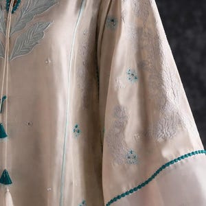 May include: Cream-colored tunic with intricate floral embroidery in light blue. The long sleeves feature delicate lace trim and small blue floral accents. A matching tassel drawstring adds a decorative touch to the neckline.