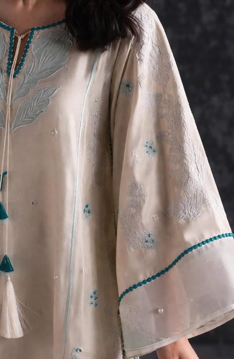 May include: Cream-colored tunic with intricate floral embroidery in light blue. The long sleeves feature delicate lace trim and small blue floral accents. A matching tassel drawstring adds a decorative touch to the neckline.