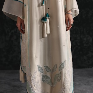 May include: Cream-colored, long-sleeved dress with intricate leaf and floral embroidery in light teal. The dress features a teal trim and tassel accents. The sleeves are wide and flowing, and the dress has a relaxed fit.