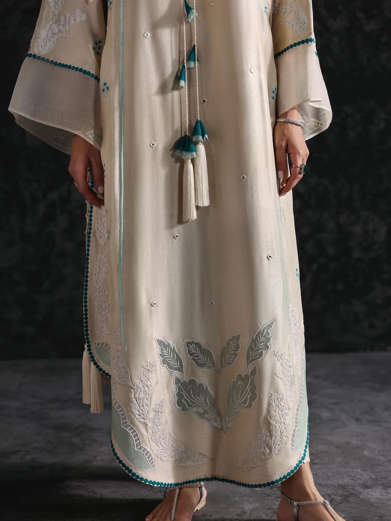 May include: Cream-colored, long-sleeved dress with intricate leaf and floral embroidery in light teal. The dress features a teal trim and tassel accents. The sleeves are wide and flowing, and the dress has a relaxed fit.