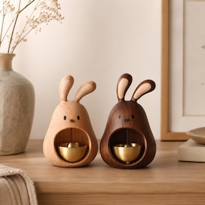 May include: Two wooden bunny-shaped ornaments. One is light-colored, the other is dark brown. Each has a gold-colored bell inside. The bunny ornaments are sitting on a wooden surface.