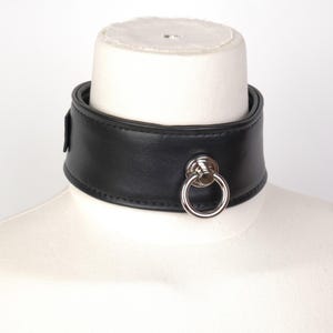 Puede incluir: Un collar negro que presenta una banda ancha y lisa con bordes cosidos y una anilla metálica de tono plateado centrada, sujeta a una base circular. El collar se muestra sobre un cuello de maniquí de color neutro sobre fondo blanco.
