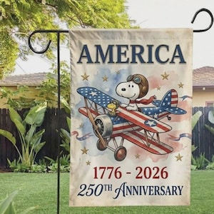 May include: A garden flag featuring Snoopy as a pilot in a red, white, and blue airplane. The flag reads "AMERICA" at the top, with "1776 - 2026 250th ANNIVERSARY" at the bottom. The background has a watercolor effect.
