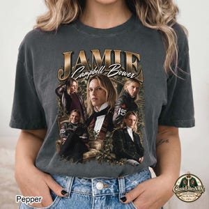 Op de afbeelding: Een antracietgrijs t-shirt met ronde hals en een grote grafische print van Jamie Campbell Bower. Het ontwerp bevat de tekst "JAMIE" en "Campbell Bower" in een gouden lettertype, omringd door vier afzonderlijke fotoportretten van de acteur.