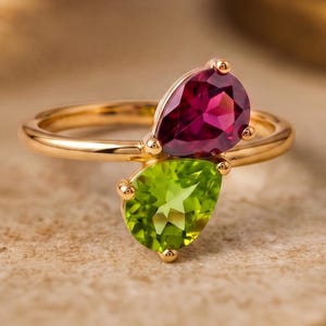 May include: A gold ring featuring two pear-shaped gemstones: a deep red garnet and a vibrant green peridot. The ring's band is a delicate gold, and the gemstones are set close together, creating a unique and elegant design. The ring is displayed on a neutral surface.