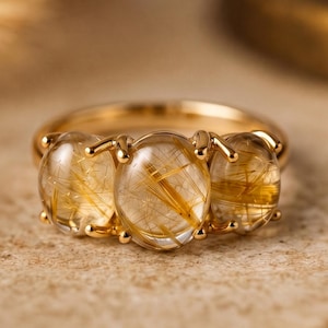 May include: A gold ring featuring three oval-cut gemstones with golden inclusions. The ring's band and settings are also gold, creating a cohesive and elegant design. The gemstones are set close together, creating a continuous look.