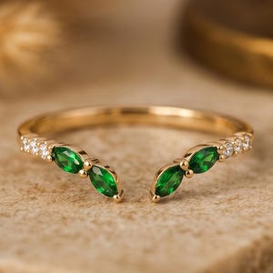 Puede incluir: Un anillo de oro con un diseño de caña dividida. El anillo presenta tres piedras preciosas verdes en forma de marquesa a cada lado, con pequeñas piedras preciosas blancas que acentúan la banda. El anillo está sobre una superficie de tonos neutros.