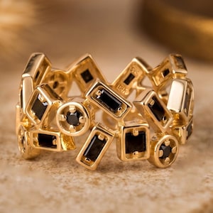 May include: A gold-colored ring with a geometric design. The ring features various shapes, including rectangles and circles, each containing a small, black stone. The ring is set on a neutral-toned surface.