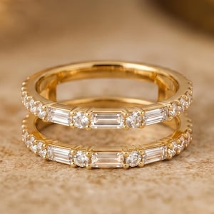 Könnte beinhalten: Zwei goldene Ringe, die übereinander liegen. Jeder Ring ist mit kleinen, klaren Edelsteinen besetzt, darunter rechteckige und runde Schliffe. Die Ringe liegen auf einem strukturierten, neutralen Hintergrund.