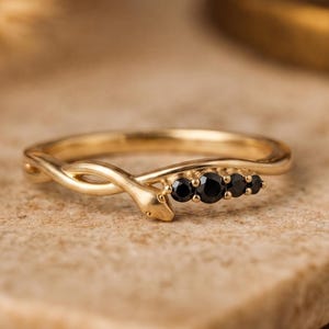 Può includere: Un anello dorato con un motivo a serpente. L'anello presenta una testa di serpente e quattro pietre nere. L'anello è appoggiato su una superficie dai toni neutri.