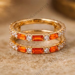 Puede incluir: Un anillo de banda dorada con un patrón alterno de piedras preciosas rectangulares de color naranja y piedras blancas redondas y transparentes. El anillo tiene un acabado metálico pulido y un diseño consistente en toda la banda.