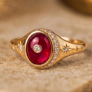 Op de afbeelding: Een gouden ring met een grote, ovale, cabochon geslepen rode edelsteen in het midden, geaccentueerd door een kleine ronde diamant. De ringband is versierd met een halve maan van kleine witte steentjes en gegraveerde stermotieven.