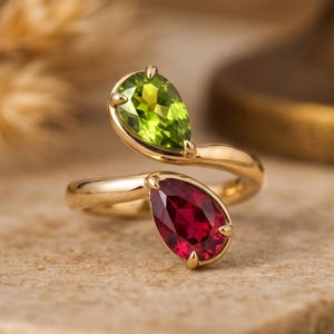 May include: A bypass ring crafted from polished gold, featuring two pear-cut gemstones. One stone is a vibrant lime green peridot, and the other is a deep red ruby. The stones are set in prong settings at the open ends of the gold band.