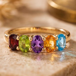 Puede incluir: Un anillo de oro con cinco piedras preciosas ovaladas en fila. Las piedras son rojas, verdes, moradas, naranjas y azules. El anillo está sobre una superficie marrón claro.