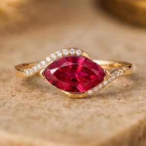 Op de afbeelding: Een gouden ring met een centrale dieprode edelsteen in marquise-slijpvorm. De band buigt rond de steen en is aan de zijkanten bezet met kleine, heldere, rond geslepen steentjes. Het metaal heeft een gepolijste afwerking en het ontwerp is symmetrisch.