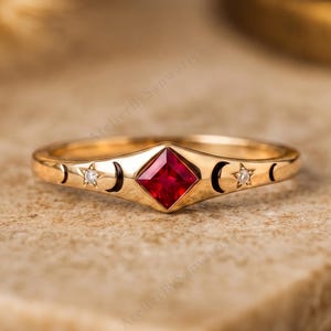 Peut inclure: Une bague en or ornée d'une pierre précieuse rouge taille princesse au centre, flanquée de deux petites pierres blanches. L'anneau est gravé de motifs célestes, incluant des formes de croissant de lune et des étoiles autour de la pierre.