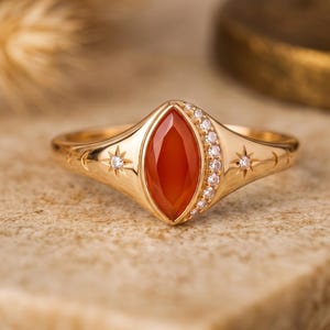May include: A gold ring featuring a central marquise-cut orange carnelian stone. The band is accented with a row of small clear stones on one side of the center gem and engraved star and crescent moon motifs on the polished gold surface.