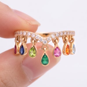 Op de afbeelding: Een gouden V-vormige ring met een rij kleine, heldere pavé-stenen langs de band. Zeven bungelende, druppelvormige edelstenen in verschillende kleuren, waaronder rood, blauw, groen, roze en oranje, hangen aan de onderkant van de ringband.