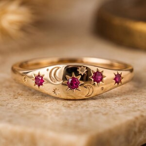 May include: A gold ring featuring a celestial motif with engraved crescent moons and stars. The band is set with four round, vibrant pinkish-red rubies embedded within star-shaped settings across the front surface of the polished metal band.