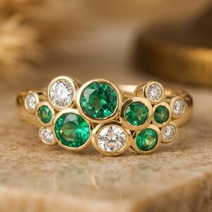 Può includere: Un anello d'oro con un design a grappolo, incastonato con smeraldi e diamanti. L'anello presenta più smeraldi e diamanti taglio rotondo, disposti molto vicini tra loro. Il design dell'anello crea un aspetto scintillante e lussuoso. L'anello è esposto su una superficie neutra.