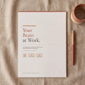 Op de afbeelding: Een planner getiteld "Your Brain at Work" met een pen en een keramische mok. De planner heeft een lichtgekleurde omslag met de titel in een groot lettertype. De pen is lichtbruin van kleur. De mok is lichtroze en wit.