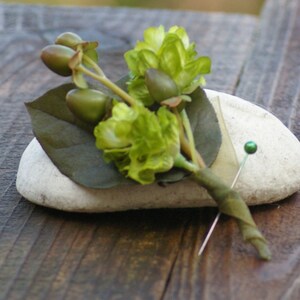 May include: A green and brown floral boutonniere with a green pin on a white rock.