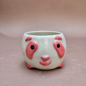 Pode incluir: Um vaso de cerâmica com rosto de panda com base creme. O panda apresenta detalhes em rosa ao redor dos olhos, orelhas e boca, com olhos pretos. O vaso é redondo e tem uma pegada pequena.